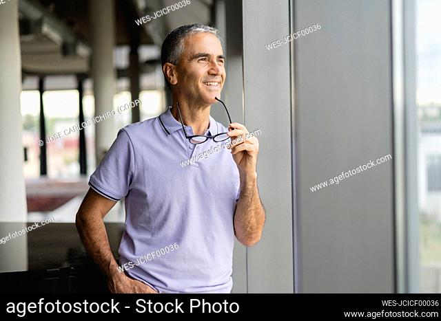 Smiling businessman holding eyeglasses looking out of window at office