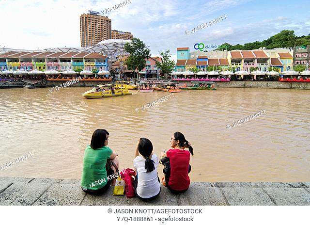 Three women sitting down on the Singapore River bank overlooking the brightly painted entertainment precinct of Clarke Quay, Singapore