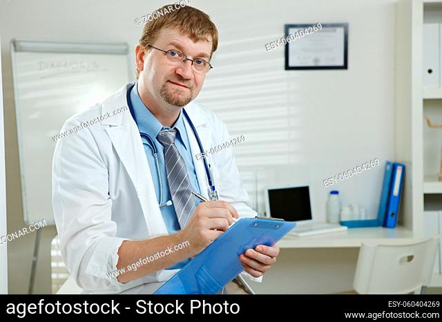 Medical office - male doctor writing on clipboard, looking up at camera