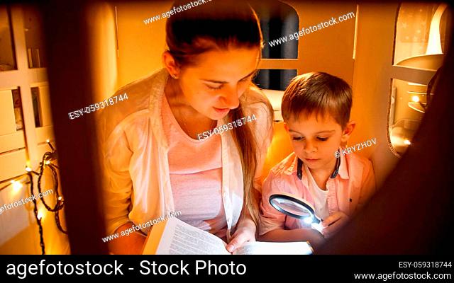 Portrait of cute boy with young mohter reading big book in tent or toy house before going to sleep. Concept of child education and family having time together...
