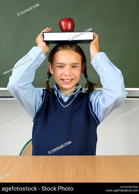 Young, smiling girl holding book on head. On book is lying apple. Looking at camera, front view