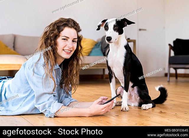 Pretty woman lying on wooden floor using a smartphone with dog sitting next to her both looking into camera