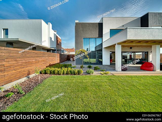 Modern country house with the large lawn and a wooden fence. In front of the house there is a covered terrace with a lounge zone