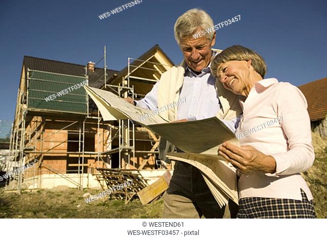 Senior couple looking at blueprints in front of incomplete built house, smiling