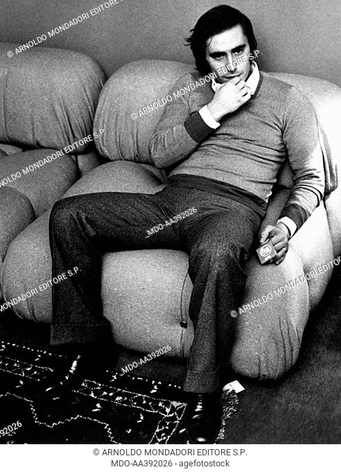 Roberto Vecchioni sitting on a sofa. Italian singer and songwriter Roberto Vecchioni sitting on sofa looking thoughtful. Milan, 1970s