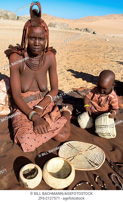 Namibia Africa remote nomadic Himba tribe young woman with child selling artwork and crafts to tourists in desert with traditional dress in desert of Hartmann...