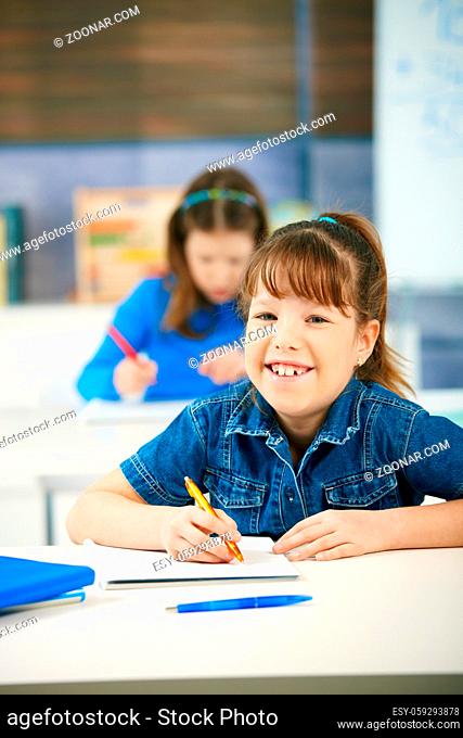 Portrait of laughing elementary age schoolgirl sitting in class looking at camera smiling, other girl in background