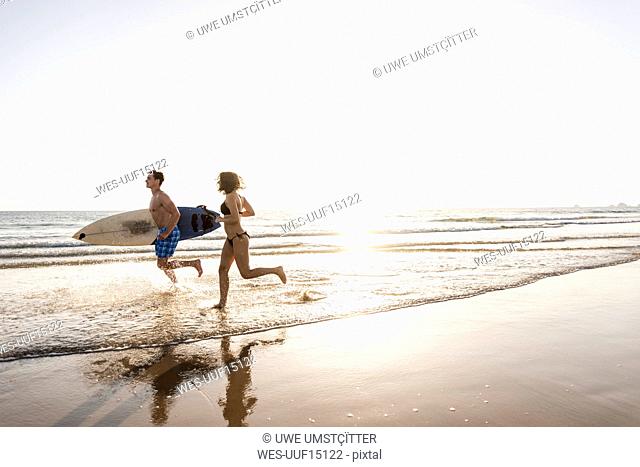 Young couple running on beach, carrying surfboard