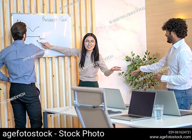 Company employee pointing with a marker at the horizontal line on the graph drawn on the whiteboard to her colleagues