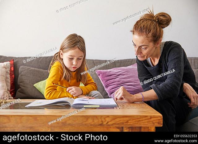 mother and her daughter, six years old girl, doing school homework with exercise book sitting on a brown sofa at home