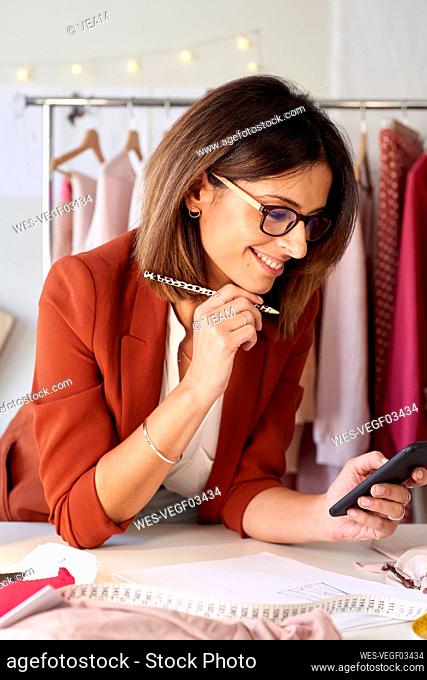Smiling fashion designer using smart phone while leaning on desk in atelier