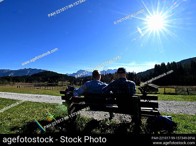 17 October 2022, Bavaria, Gerold: Two vacationers from Hamburg enjoy the sun on a bench at Geroldsee, the Karwendel mountains can be seen in the background