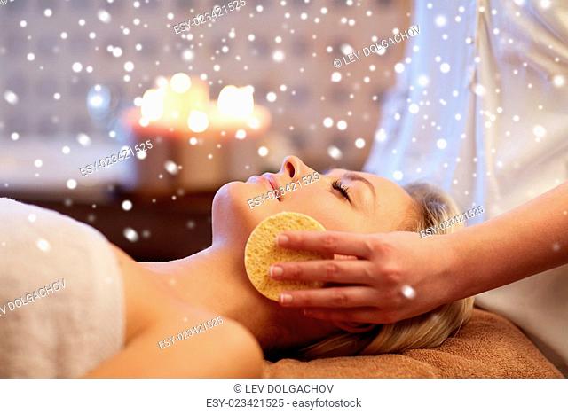 people, beauty, spa, skin care and relaxation concept - close up of beautiful young woman lying with closed eyes and having face massage with sponge in spa with...