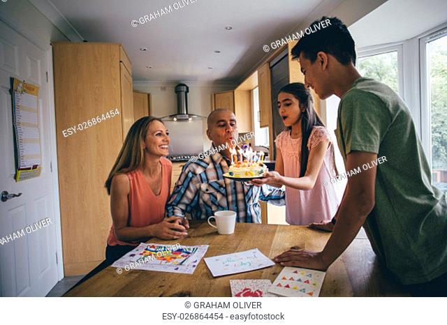Family of four celebrating the dads birthday. His daughter is holding a cake they have made and is helping him blow out the candles