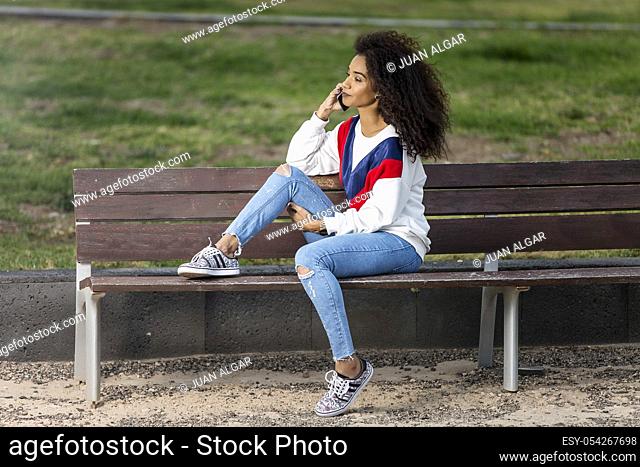 Stylish ethnic model in jeans and bright hoodie speaking on smartphone while chilling on bench in park