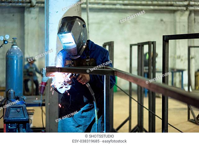 front close-up of a worker wearing protective gear, with mask and a leather apron, welding together two pieces of square pipes into a frame