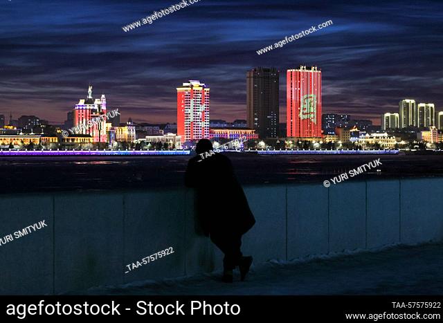 RUSSIA, BLAGOVESHCHENSK - FEBRUARY 25, 2023: A pedestrian on the promenade by the Amur River enjoys a view of the Chinese city of Heihe at dusk in winter