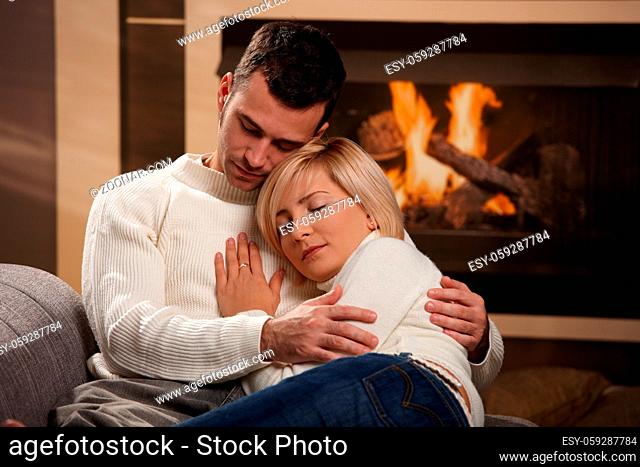 Young couple hugging on sofa in front of fireplace at home, eyes closed