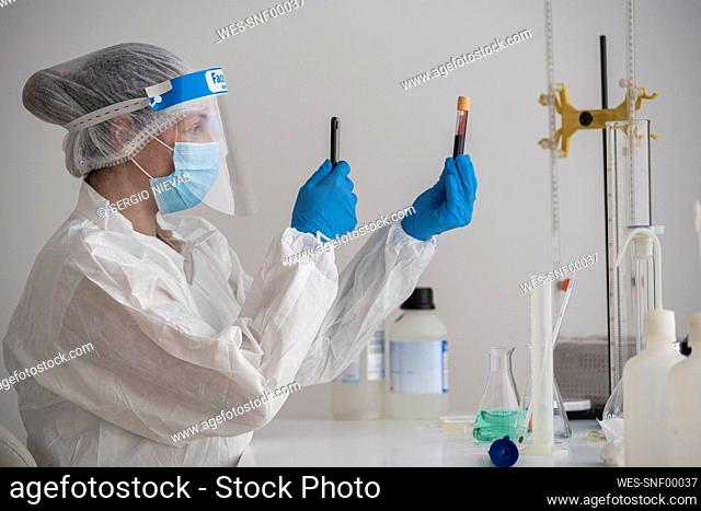 Woman in protective wear sitting at desk holding smartphone and blood sample