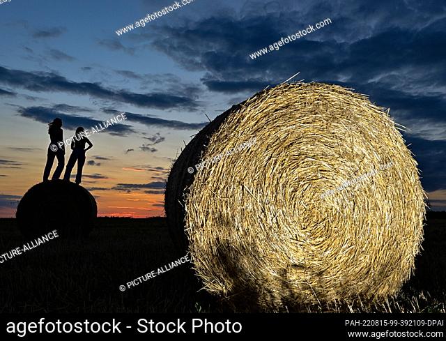 08 August 2022, Brandenburg, Jacobsdorf: Two girlfriends are standing on a roll of straw in a field at sunset. Photo: Patrick Pleul/dpa