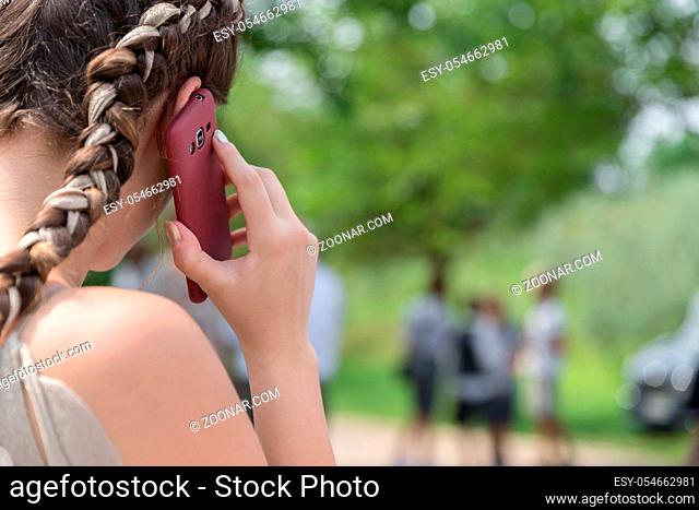 Close-up over the shoulder young girl talking on the mobile phone with green foliage and blurry people on background