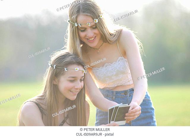 Two teenage girls wearing daisy chain headdresses looking at instant photographs in park