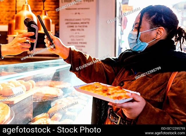 Italy, Woman in face mask standing paying with smart phone for snack on street