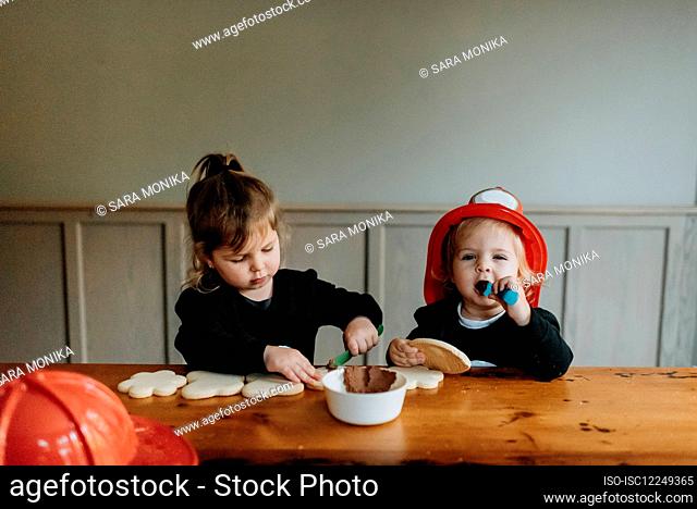 Two children dressed as firefighters at a table decorating cookies with chocolates and sprinkles