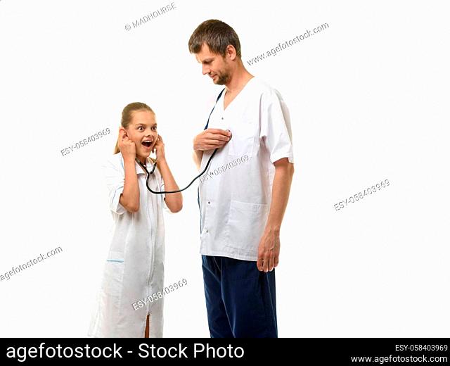 The doctor put the head of the phonendoscope to his heart, a girl in a medical coat listens with surprise to the doctor's heart