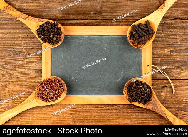 Four wooden cooking spoons made of olive wood with different peppercorns and an old black slate board with copy space on a rustic wooden background
