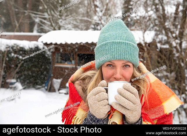 Mature woman wearing knit hat drinking coffee at backyard during snow