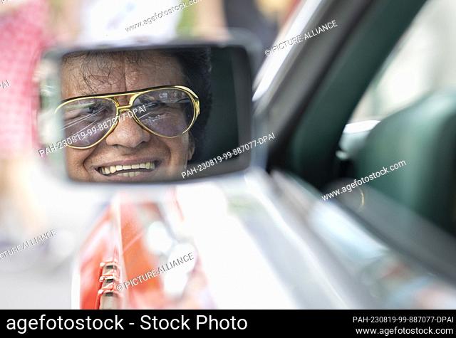 19 August 2023, Hesse, Bad Nauheim: Styled as Elvis, 70-year-old Lello Marino looks into the rearview mirror of his U.S. road cruiser at the Elvis Festival