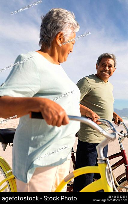Happy senior multiracial couple looking at each other while standing with bicycles on sunny beach