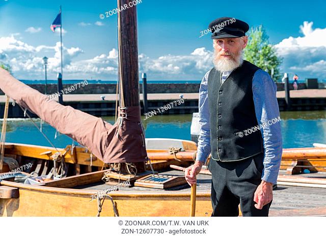 Urk, The Netherlands - September 02, 2017: Old sailor with stick, cap and beard standing for traditional wooden fishing boat in Dutch harbor of Urk