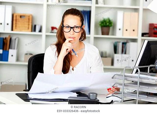Serious young business woman wearing glasses working at her desk in the office holding a spreadsheet in her hands