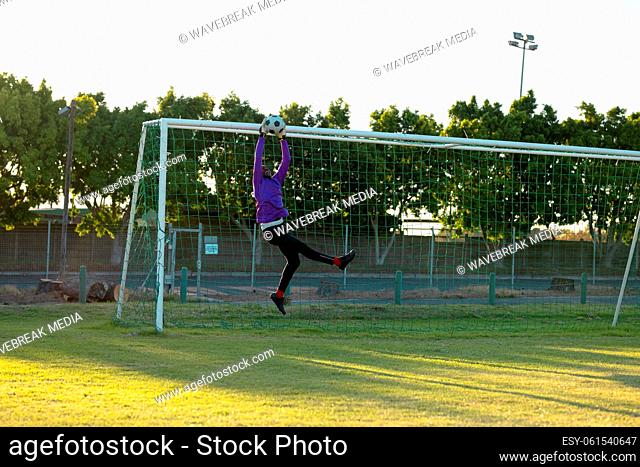 African american male goalkeeper with arms raised jumping and catching soccer ball in mid-air