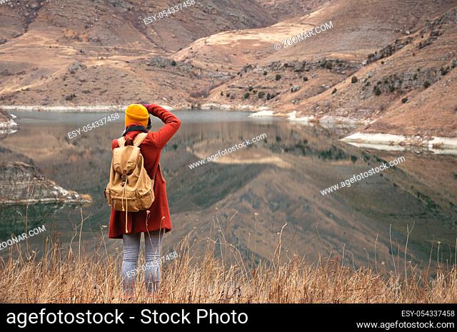 A portrait from the back of a girl traveler take a picture on the background of a lake in the mountains in autumn or early spring. Travel concept