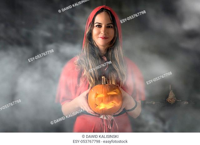 Halloween portrait, adult Caucasian white woman holding orange pumpkin, dressed and makeup stylised for Red Riding Hood on dark smoky background