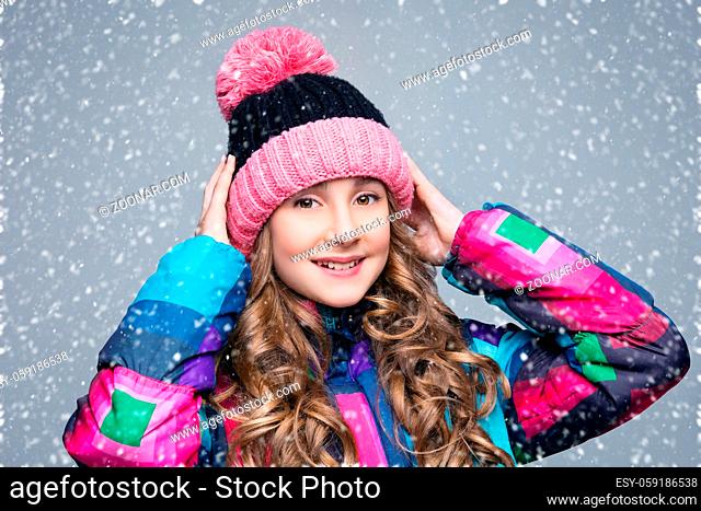 Beautiful happy teen girl with long curly hair in pink wool hat and bright warm coat. Studio shot over grey background with falling snow. Copy space