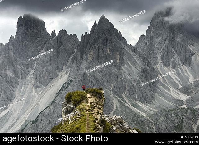 Man with red jacket standing on one degree, behind him mountain peaks and pointed rocks, dramatic clouds, Cimon the Croda Liscia and Cadini group