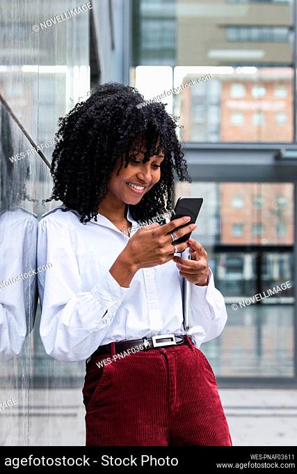 Smiling businesswoman using mobile phone leaning on wall