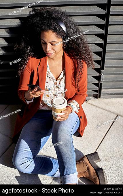 Young woman using smart phone holding coffee cup while sitting on footpath