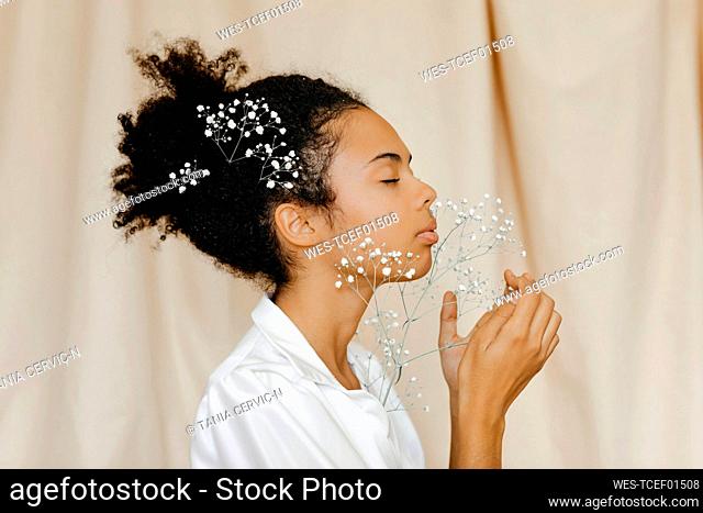 Woman with eyes closed smelling white flowers by beige curtain