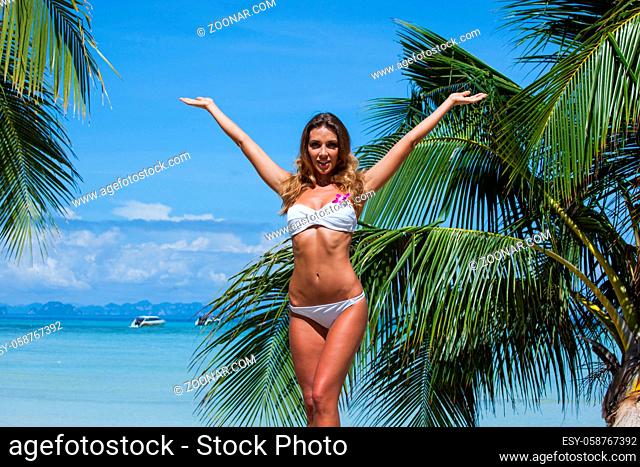 Portrait of young model woman in bikini raising her arms at tropical beach with palms in Thailand