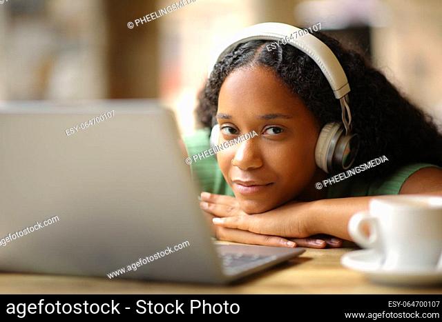 Black woman with headphone and laptop looks at you in a coffee shop terrace