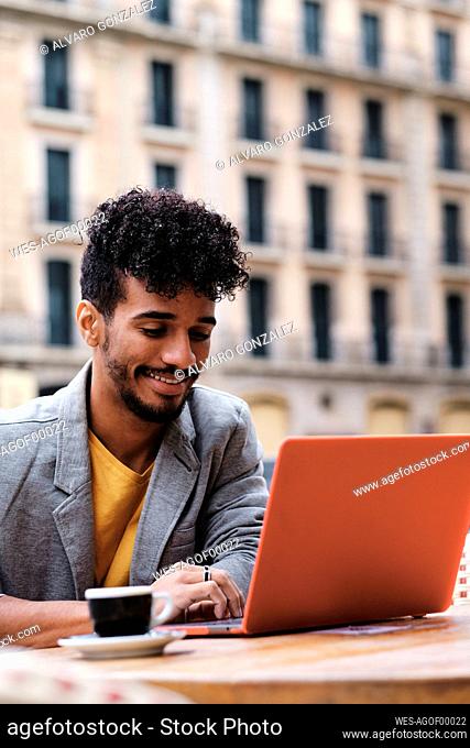 Fashionable man smiling while working on laptop sitting at side walk cafe
