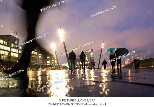 05 November 2019, Hamburg: Passers-by are on their way along the promenade at the landing stages in light rain and dusk. In the background you can see the...