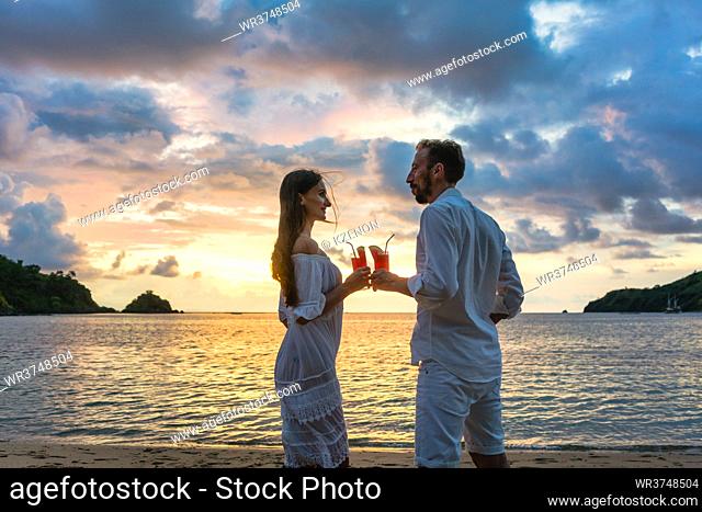 Couple in tropical vacation with drinks toasting on beach by the sea during colorful sunset