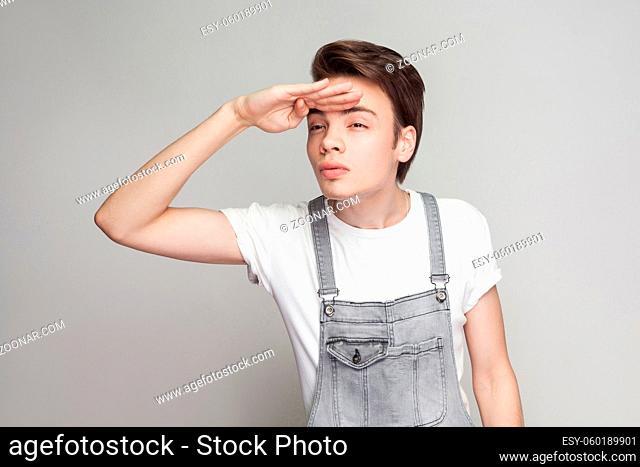 Serious brunette man in casual style with white t-shirt and denim overalls standing holding hands on forehead and looking to far with serious face