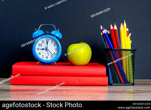 The blue alarm clock along with multi-colored pencils stand on a stack of red books, next to an apple